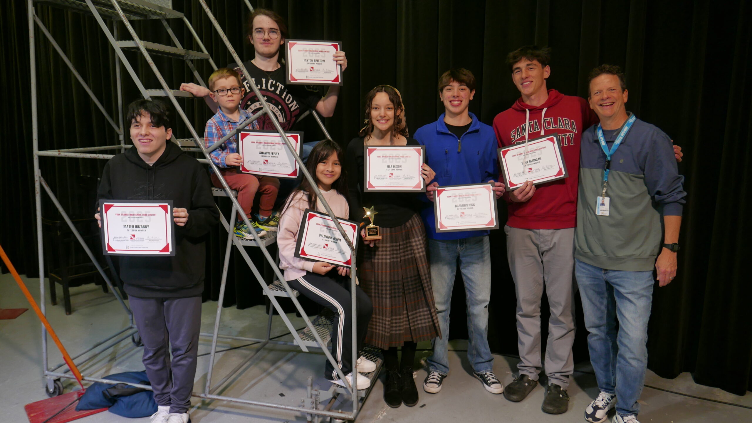Students sit together on stairs with their certificates from winning the video contest.